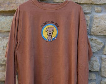 May include: A long-sleeved, brown t-shirt hanging on a white hanger. The shirt features a cartoon dog graphic inside a blue and yellow circle, with the words "Diggin' my dog" above the dog.
