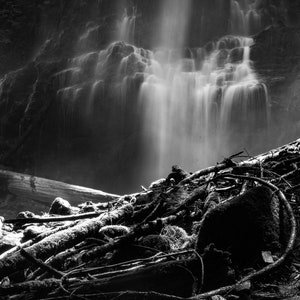 May include: A black and white photograph of a waterfall cascading over a rocky cliff. The water is flowing over the rocks and creating a mist. The bottom of the image shows a pile of fallen branches and logs.