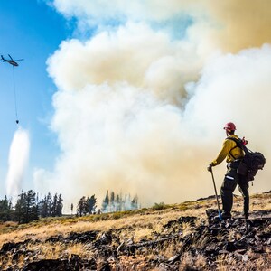 May include: A firefighter in a yellow jacket and red helmet stands on a rocky hillside, looking up at a helicopter dropping water from a bucket to extinguish a wildfire. Smoke billows in the background.