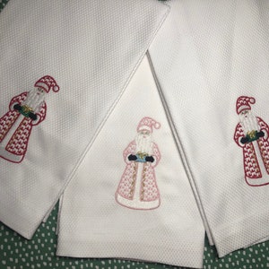 May include: Three white kitchen towels with embroidered images of Santa Claus holding a gift. The Santa Claus on the left towel is red, the middle towel is pink, and the Santa Claus on the right towel is red.