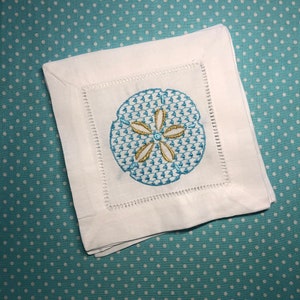 May include: White linen cocktail napkins with a blue and gold embroidered sand dollar design. The napkins are square with a decorative stitched border. The background is a light blue fabric with white polka dots.