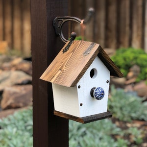 May include: A handcrafted wooden birdhouse with a white body and a brown, textured roof. The birdhouse features a round entry hole and a decorative blue and white knob. It is mounted on a dark brown post with a metal hook.