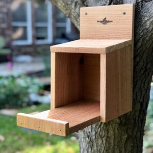 May include: A wooden birdhouse with a small opening and a pull-out tray. The birdhouse is mounted on a tree trunk and has the text "The Birdhouse Factory" on the front.