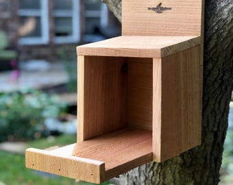 Può includere: Una casetta per uccelli in legno con una piccola apertura e un vassoio estraibile. La casetta per uccelli è montata su un tronco d'albero e ha il testo "The Birdhouse Factory" sul davanti.