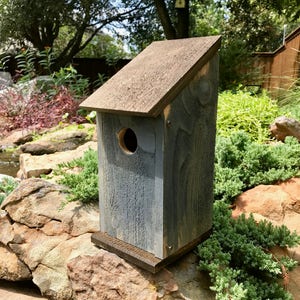 Puede incluir: Una casa para p&aacute;jaros de madera con un acabado gris desgastado y un techo inclinado de color marr&oacute;n oscuro. La estructura presenta un &uacute;nico orificio de entrada circular en el panel frontal y se asienta sobre una base marr&oacute;n oscuro.