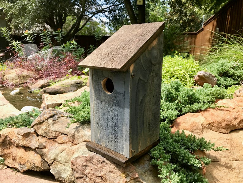Puede incluir: Una casa para p&aacute;jaros de madera con un acabado gris desgastado y un techo inclinado de color marr&oacute;n oscuro. La estructura presenta un &uacute;nico orificio de entrada circular en el panel frontal y se asienta sobre una base marr&oacute;n oscuro.