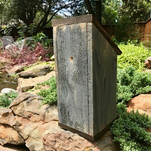 Puede incluir: Una casa para p&aacute;jaros alta y rectangular construida con madera desgastada de tono gris y un techo inclinado. El panel frontal presenta un &uacute;nico orificio de entrada circular peque&ntilde;o situado cerca del centro superior, con una textura de veta natural.