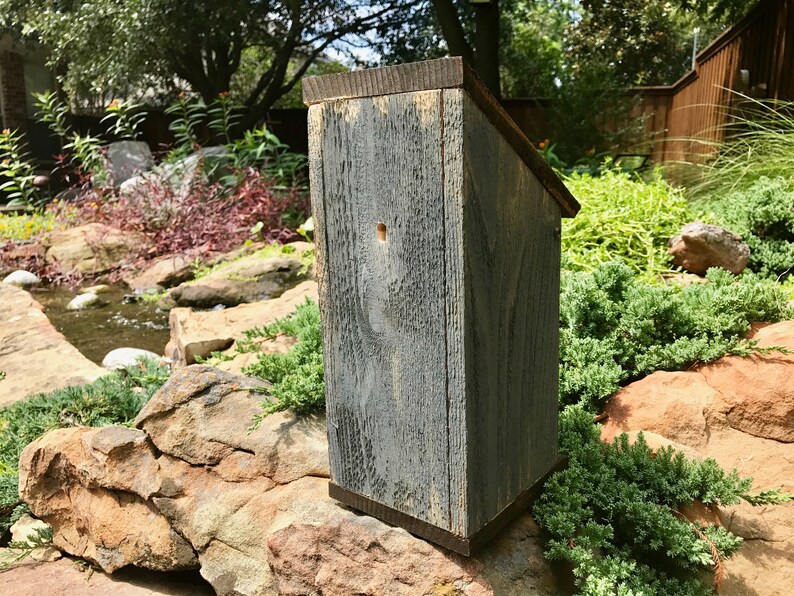 Puede incluir: Una casa para p&aacute;jaros alta y rectangular construida con madera desgastada de tono gris y un techo inclinado. El panel frontal presenta un &uacute;nico orificio de entrada circular peque&ntilde;o situado cerca del centro superior, con una textura de veta natural.