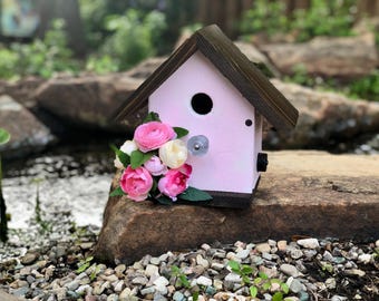 Può includere: Una casetta per uccelli rosa con un tetto marrone e un piccolo foro per permettere agli uccelli di entrare. La casetta è decorata con un bouquet di fiori rosa e bianchi.