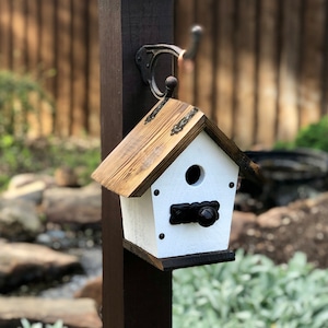 Puede incluir: Una casita de pájaros blanca con un techo de madera marrón y detalles metálicos negros. La casita tiene una pequeña abertura para que los pájaros entren y una percha en la parte delantera.