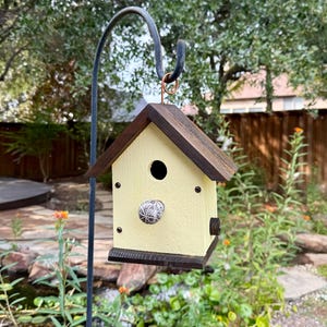 May include: A yellow birdhouse with a dark brown roof and trim hangs from a black metal hook. The birdhouse has a round entry hole and a decorative knob. The background includes a garden setting with greenery and a pond.