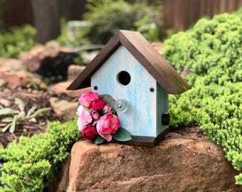 Può includere: Una casetta per uccelli blu chiaro con un tetto marrone e un piccolo foro per l'ingresso degli uccelli. La casetta è decorata con un pomello di vetro e fiori rosa.