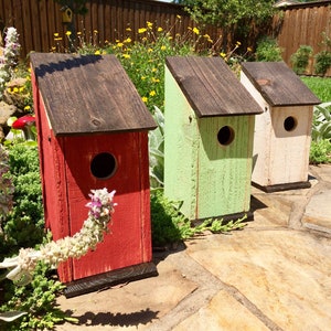 Puede incluir: Tres casas para p&aacute;jaros de madera con techos inclinados y orificios de entrada circulares. La primera est&aacute; pintada de rojo, la segunda de verde claro y la tercera de color blanco roto. Cada casa tiene techo y base marr&oacute;n oscuro con textura.
