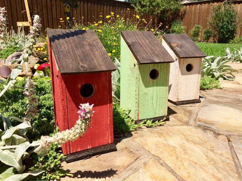 Puede incluir: Tres casas para p&aacute;jaros de madera con techos inclinados y orificios de entrada circulares. La primera est&aacute; pintada de rojo, la segunda de verde claro y la tercera de color blanco roto. Cada casa tiene techo y base marr&oacute;n oscuro con textura.