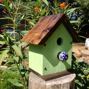 May include: A green birdhouse with a brown roof and a blue and white ceramic knob. The birdhouse is mounted on a wooden stump.