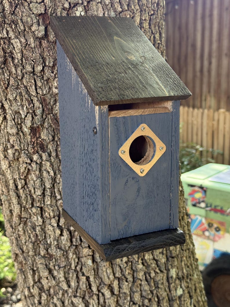 Puede incluir: Una casa para p&aacute;jaros de madera con cuerpo pintado en azul oscuro, techo inclinado marr&oacute;n oscuro y base marr&oacute;n oscuro. El panel frontal presenta un orificio de entrada circular rodeado por una placa met&aacute;lica cuadrada clara con cuatro tornillos.