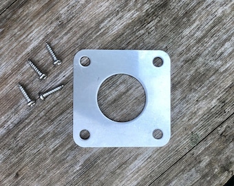 Può includere: Una piastra metallica quadrata color argento con un foro circolare centrale e quattro fori agli angoli. Quattro viti argentate sono sparse nelle vicinanze. La piastra è probabilmente una staffa di montaggio o un componente hardware simile.