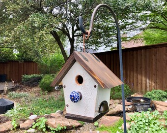 May include: A white birdhouse with a brown roof and a blue and white ceramic knob on the front. The birdhouse is hanging from a metal hook.