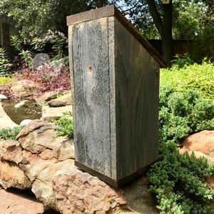 Puede incluir: Una casa para p&aacute;jaros vertical de madera con un acabado gris desgastado y un techo inclinado. La estructura presenta un peque&ntilde;o orificio de entrada circular en el panel frontal, mostrando una veta de madera rugosa y texturizada en el exterior.