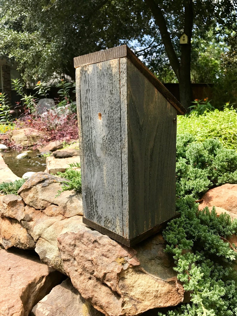 Puede incluir: Una casa para p&aacute;jaros vertical de madera con un acabado gris desgastado y un techo inclinado. La estructura presenta un peque&ntilde;o orificio de entrada circular en el panel frontal, mostrando una veta de madera rugosa y texturizada en el exterior.