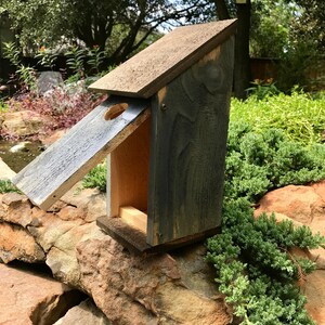 Puede incluir: Una casa para p&aacute;jaros de madera con techo inclinado y acabado te&ntilde;ido en gris oscuro. La estructura presenta un &uacute;nico orificio de entrada circular en el panel frontal, textura de veta de madera visible y una base rectangular, ensamblada con tornillos met&aacute;licos visibles.