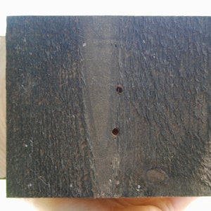 Puede incluir: Una pieza rectangular de madera te&ntilde;ida de marr&oacute;n oscuro con una superficie de grano rugoso y texturizado. La madera presenta dos peque&ntilde;os orificios circulares perforados verticalmente en el centro, revelando el tono natural m&aacute;s claro en el interior.