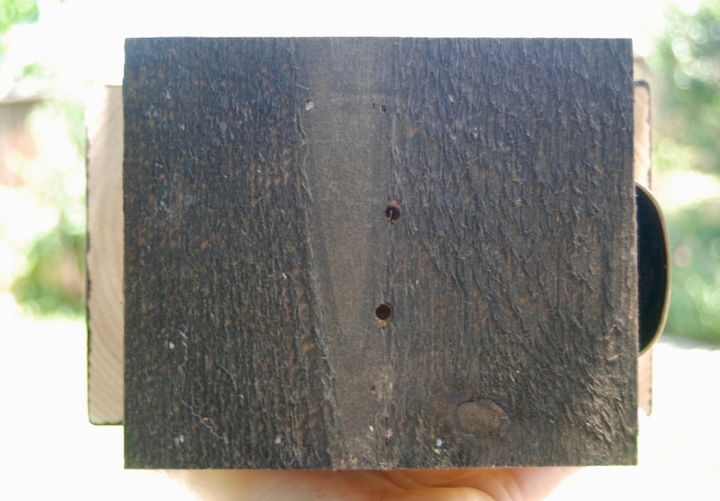 Puede incluir: Una pieza rectangular de madera te&ntilde;ida de marr&oacute;n oscuro con una superficie de grano rugoso y texturizado. La madera presenta dos peque&ntilde;os orificios circulares perforados verticalmente en el centro, revelando el tono natural m&aacute;s claro en el interior.