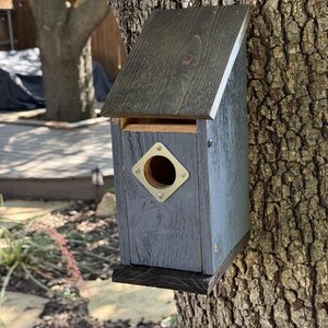 Puede incluir: Una casa para p&aacute;jaros de madera con un techo inclinado te&ntilde;ido de oscuro y un cuerpo pintado de gris. El frente presenta una placa de metal cuadrada con un orificio de entrada circular asegurado por cuatro tornillos. Montada sobre un tronco.