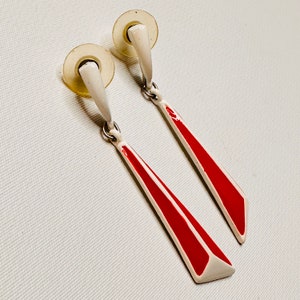 May include: A pair of red and white clip-on earrings with a geometric design. The earrings are made of metal and have a white base with a red stripe down the center.
