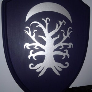 Puede incluir: Un escudo de madera de color morado oscuro con una luna creciente plateada y un diseño de árbol plateado estilizado.