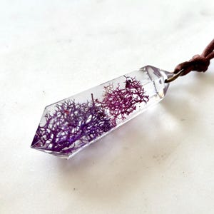 May include: A clear resin pendant with a geometric shape, showcasing vibrant purple and red plant life encased within. The pendant hangs from a brown cord, creating a unique and naturalistic necklace.