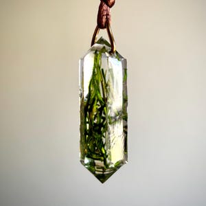 May include: A clear, crystal-shaped pendant with green plant material encased inside. The pendant is suspended from a brown cord and a gold-colored metal ring. The background is a soft, neutral color.