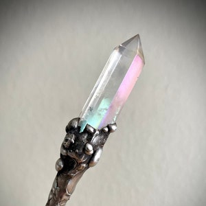 May include: A clear quartz crystal wand with iridescent colouring at the tip. The wand is held by a detailed silver-coloured metal handle with an ornate design. The crystal is approximately 15 cm long.