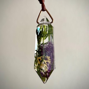May include: A clear, prism-shaped pendant with preserved greenery and purple moss encased in resin. The pendant hangs from a brown cord and a copper-colored metal loop. The design features a natural, botanical theme.