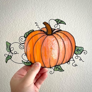 May include: A stained glass pumpkin with a black metal frame and green leaves. The pumpkin is orange with a light orange center.