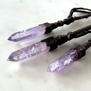Könnte beinhalten: Drei Amethyst-Kristallanhänger mit dunkelbraunen Lederkordeln. Die Kristalle sind helllila und in dunklem, strukturiertem Metall eingefasst. Die Anhänger werden vor einem weißen Hintergrund präsentiert.