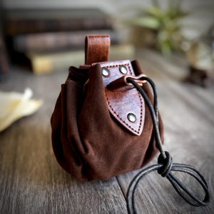 May include: A brown leather pouch with a drawstring closure and a flap with a leather strap and metal rivets. The pouch is sitting on a wooden surface.
