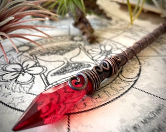 May include: A red crystal wand with copper wire wrapped around the handle. The wand is lying on a white surface with a black and white design.