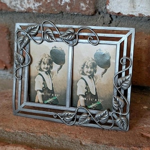 May include: A silver-colored metal double picture frame with a leaf and vine design. The frame holds two vintage photos of a child. The photo size is 1.5" x 2.5". The frame is displayed on a brick surface.