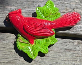 May include: A vibrant red cardinal bird sculpture perched on green leaves. The bird has detailed feather textures and a prominent crest. The leaves are a bright green, creating a striking contrast against the red bird.