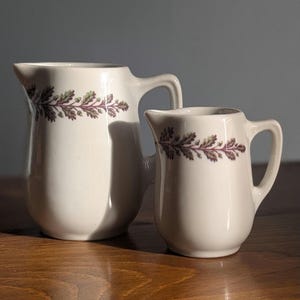 May include: Two white ceramic pitchers with a decorative leaf pattern in green and purple. The pitchers have a classic design with a handle and spout, suitable for serving beverages or displaying flowers. The pitchers are of different sizes.