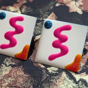 May include: Two square earrings with a white base. Each earring features a bright pink squiggly line, a blue glitter dot, and an orange glitter accent. The earrings are set against a patterned background.