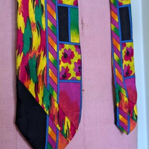 May include: A colorful silk tie with a geometric pattern featuring pink flowers, yellow, green, orange, and blue stripes. The tie has a black background.