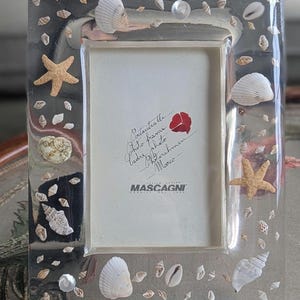May include: Clear rectangular photo frame decorated with seashells, starfish, and small clear beads. The frame has a white interior and the brand name "Mascagni" is visible. The frame is a decorative item.