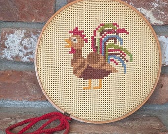 May include: A round, cross-stitched rooster design in a wooden hoop. The rooster features brown, red, green, and blue details on a beige background. A red cord is attached to the hoop. The artwork is displayed against a brick wall.