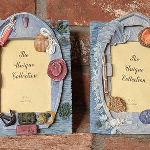 May include: Two decorative picture frames with a nautical theme. Each frame is shaped like a boat and features fishing-related embellishments, including a hat, fishing rod, and tackle. The frames have the text "The Unique Collection".