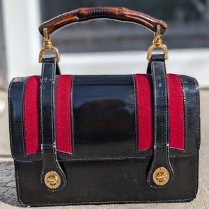 May include: Black patent leather handbag with red and black accents. The bag features a bamboo handle and gold-tone hardware. The design includes red fabric stripes down the front. The bag has a rectangular shape and a clasp closure.
