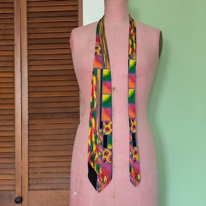 May include: A colorful patterned tie with geometric shapes in shades of yellow, green, orange, pink, and black. The tie is displayed on a pink dress form.