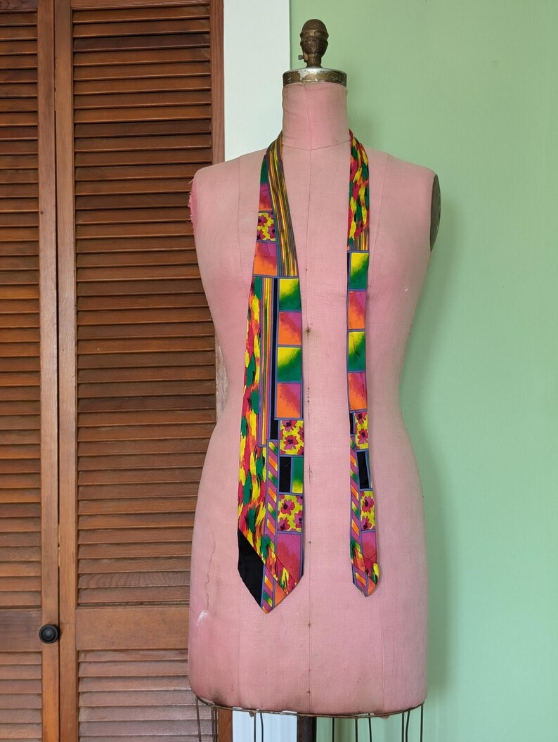 May include: A colorful patterned tie with geometric shapes in shades of yellow, green, orange, pink, and black. The tie is displayed on a pink dress form.
