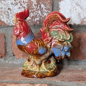 May include: A ceramic rooster figurine with a red comb and wattle, blue and brown body, and green and brown tail feathers. The rooster is perched on a base with a floral design. The figurine is a decorative item.